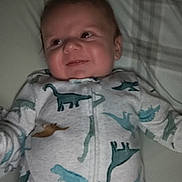 Braxton is registered to the contest to win money with this photo: baby, infant, smile, dinosaur_print, onesie, bed, pillow, blanket, clothing, child, happy, face, lying_down, indoors, cute, young, person, soft_lighting, sleepwear, comfortable