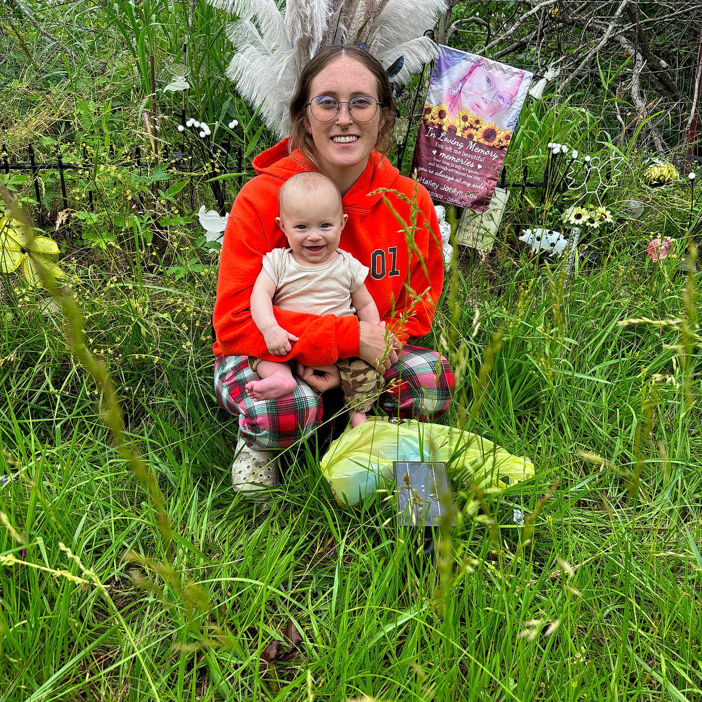 Creed Steele Fischer is registered to the contest to win money with this photo: baby, child, crouching, decorations, feathers, fence, glasses, grass, greenery, happy, memorial, nature, orange_hoodie, outdoor, person, plaid_pants, plants, portrait, smile, woman