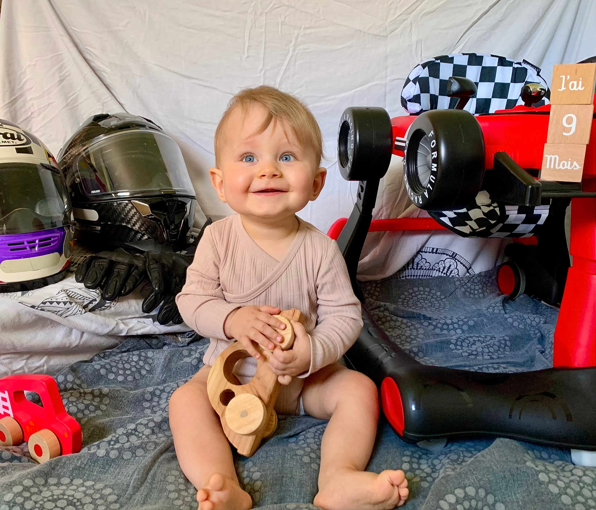 Caleb participe au concours pour gagner de l'argent avec cette photo : baby, infant, blue_eyes, wooden_toy, toy_car, motorcycle_helmet, motorcycle_gloves, red_toy_car, checkered_flag, wooden_blocks, blanket, smiling, sitting, indoors, child, cute, playtime, milestone, happy, portrait