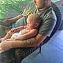 Brodi is registered to the contest to win money with this photo: baby, beard, casual_clothing, daylight, diaper, greenery, holding, man, outdoor, peaceful, phone, porch, portrait, relaxation, short_hair, sitting, sleeping, tattoo, wicker_chair, wooden_floor