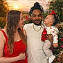 baby, casual_clothing, family, flowers, garden, gold_chain, happy, headband, holding, joyful, man, nature, outdoor, people, portrait, red_dress, smile, sunset, white_shirt, woman