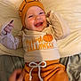 baby, child, smiling, halloween, pumpkin, hat, orange_clothing, beige_clothing, bed, blanket, cute, happy, infant, lying_down, indoor, face, person, celebration, festive, holiday