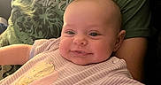 Valerie is registered to the contest to win money with this photo: baby, infant, child, smiling, portrait, indoors, caregiver, green_shirt, onesie, clothing, sitting, lying_down, close_up, face, eyes, warm_lighting, window, couch, home, family