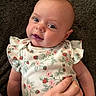 baby, infant, child, portrait, close_up, smiling, happy, face, eyes, floral_clothing, strawberry_pattern, ruffled_sleeves, lying_down, indoor, carpet, hand, parent, caregiver, rosy_cheeks, adorable