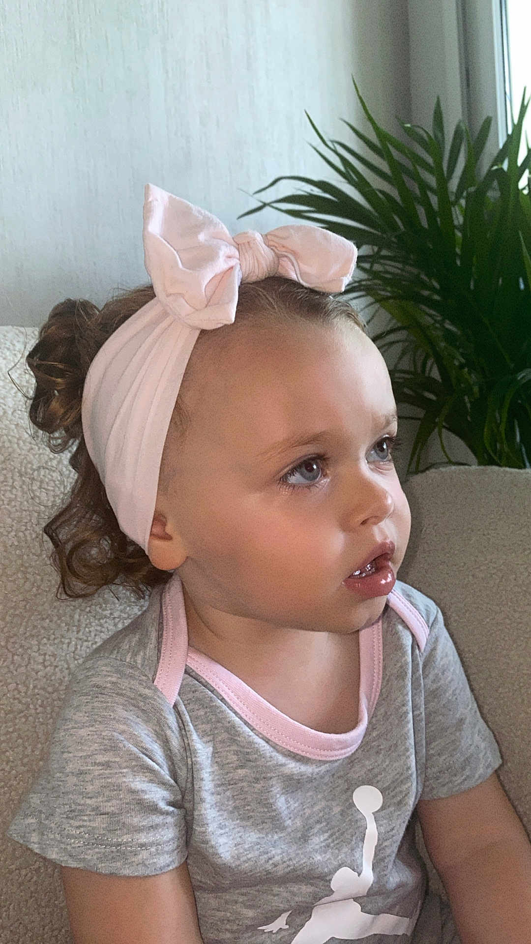 Inaya a rejoint le concours — aidez-le/la à gagner de superbes lots ! toddler, child, headband, pink_bow, curly_hair, gray_shirt, indoor, plant, greenery, chair, portrait, cute, young_child, sitting, casual_clothing, closeup, face, looking_away, home, soft_lighting