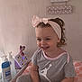 toddler, child, smiling, headband, bow, grey_clothing, sitting, baby_care_products, toy, purple_dog_toy, changing_pad, floral_pattern, bracelet, wall_decor, indoor, happy, person, cute, young_child, casual