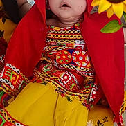 Kiarah is registered to the contest to win money with this photo: baby, child, traditional_clothing, red_shawl, yellow_skirt, sunflower, face, person, sitting, colorful, patterned_fabric, indoor, hand, flower, headwear, cute, expression, bright_colors, textile, human