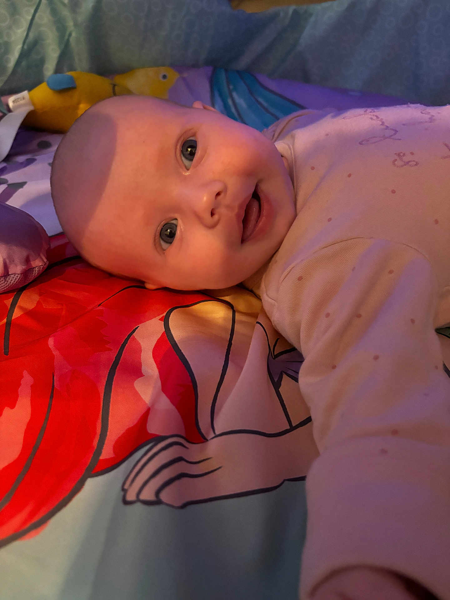 Lilith is registered to the contest to win money with this photo: baby, infant, child, face, smile, bedspread, colorful, plush_toy, bird_toy, cartoon, fabric, sleepwear, person, portrait, indoors, soft_light, cute, young, laying_down, closeup