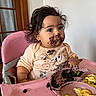 baby, baby_clothing, child, chocolate, dark_hair, earring, feeding, food, food_tray, high_chair, home, indoor, mealtime, messy_face, pink_chair, portrait, scrambled_eggs, sitting, smiling, toddler