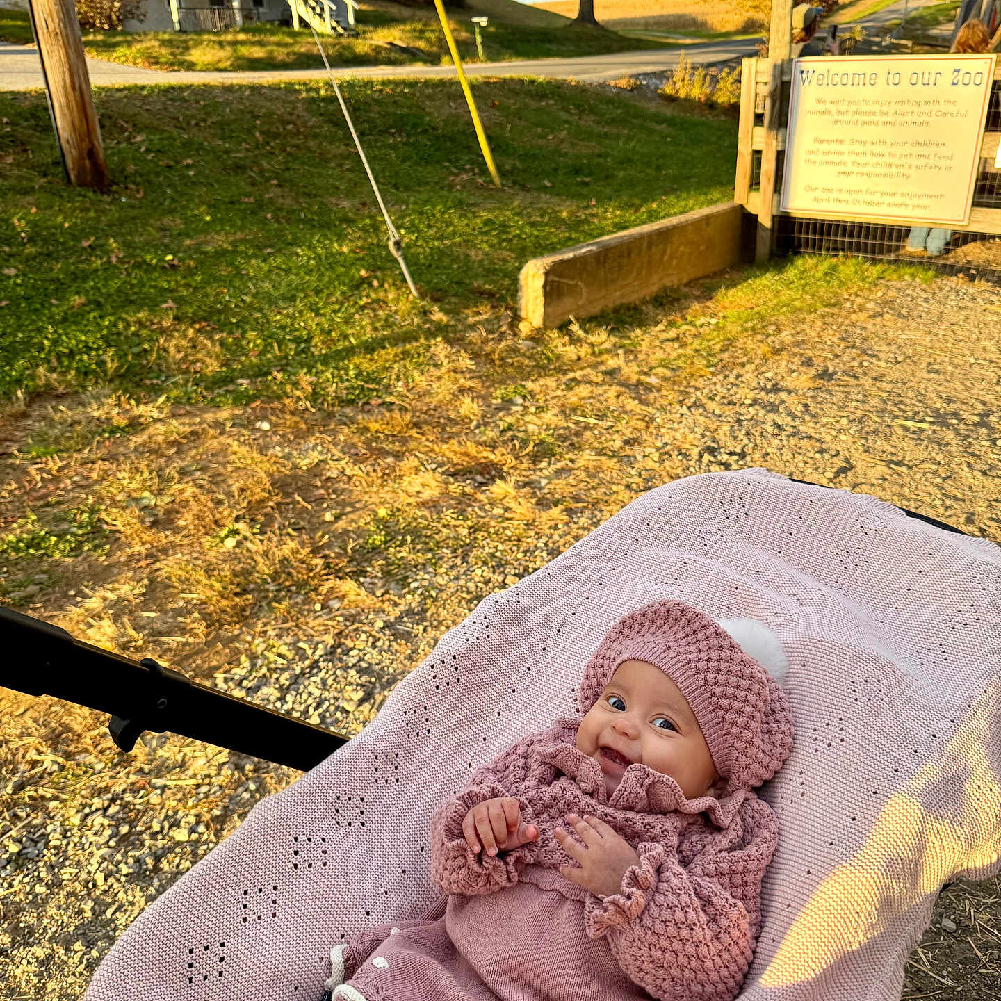 Mía Issabela is registered to the contest to win money with this photo: baby, blanket, child, daylight, fence, grass, happy, house, knit_hat, knit_sweater, nature, outdoor, park, pink_clothing, road, sign, smile, stroller, sunlight, tree