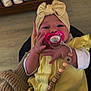 baby, pacifier, headband, yellow_clothing, hand, crib, blanket, wooden_floor, pacifier_clip, beads, toddler, indoors, person, child, cute, sitting, knit_blanket, infant, face, clothing