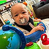 Carson is registered to the contest to win money with this photo: baby, pacifier, activity_center, toy, living_room, sofa, dog, child, infant, face, hand, colorful, furniture, indoor, play, person, comfort, cute, home, pet