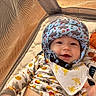 Carson is registered to the contest to win money with this photo: baby, helmet, bib, smile, playpen, mesh, blanket, plush_toy, infant, cute, child, clothing, patterned, indoor, soft, padding, happy, face, person, toy