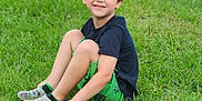 Logan is registered to the contest to win money with this photo: child, boy, grass, smiling, outdoor, greenery, casual_clothing, shorts, tshirt, shoes, people, spectators, baseball, sports, field, fence, chair, game, fun, daytime
