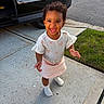 child, toddler, smiling, happy, outdoor, driveway, sidewalk, grass, car, portrait, standing, curly_hair, dress, skirt, socks, shoes, daylight, suburban, person, face