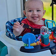 Dean is registered to the contest to win money with this photo: baby, child, smiling, tongue_out, walker, toys, colorful, indoor, happy, playing, person, infant, red_clothing, blue, plastic, seat, face, hand, closeup, cute