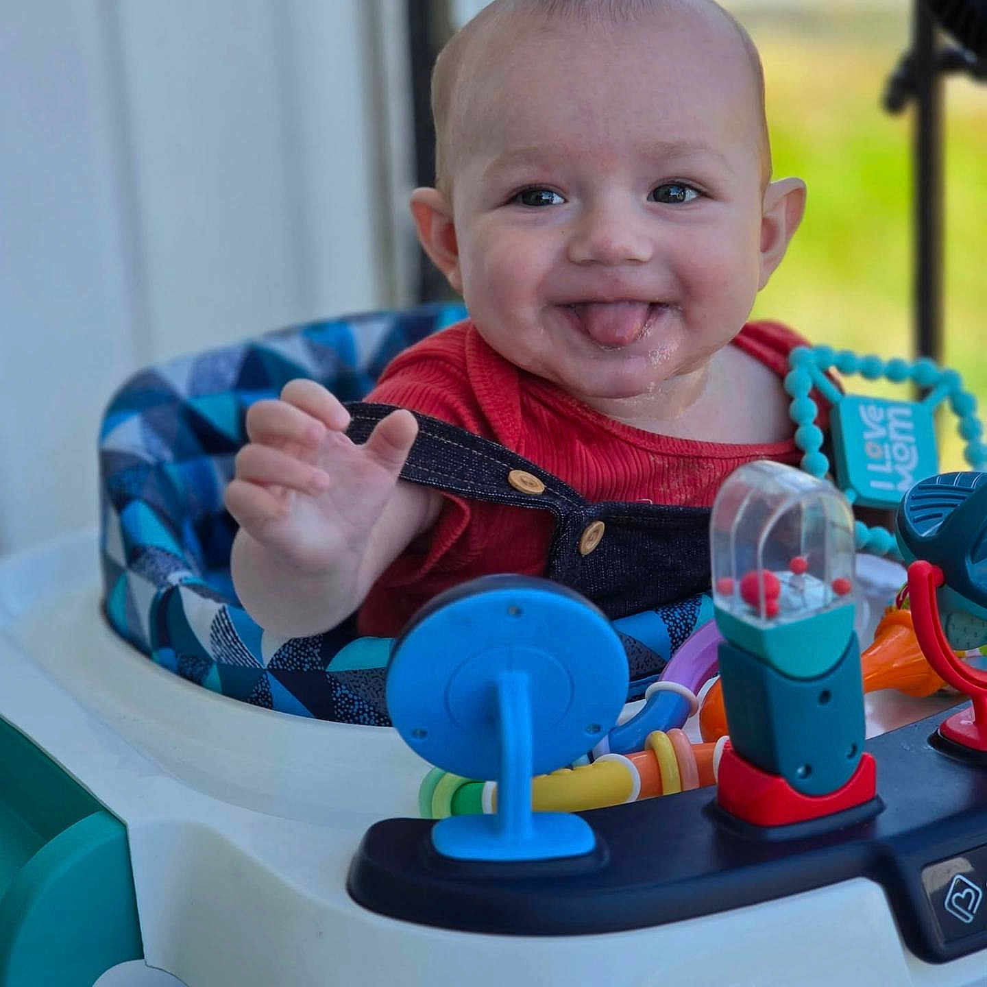 Dean is registered to the contest to win money with this photo: baby, child, smiling, tongue_out, walker, toys, colorful, indoor, happy, playing, person, infant, red_clothing, blue, plastic, seat, face, hand, closeup, cute