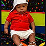 baby, child, red_shirt, white_shorts, white_cap, brown_shoes, carpet, colorful_background, sitting, serious_expression, indoor, portrait, footwear, hat, person, patterned_floor, cute, fashion, casual_clothing, infant