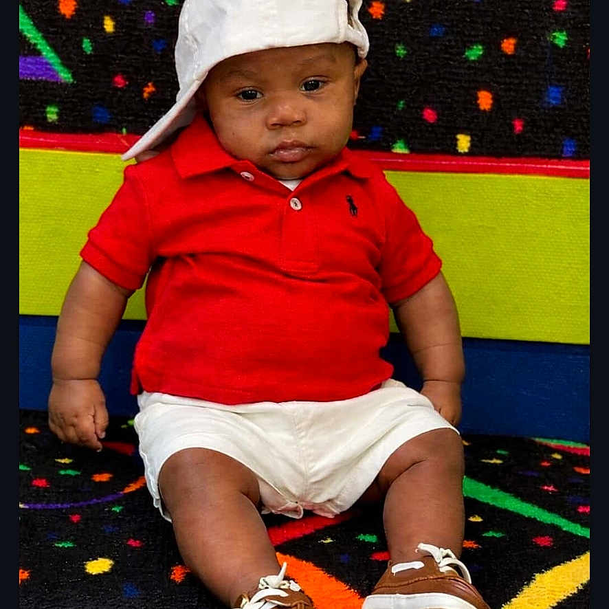Cam'Ron is registered to the contest to win money with this photo: baby, child, red_shirt, white_shorts, white_cap, brown_shoes, carpet, colorful_background, sitting, serious_expression, indoor, portrait, footwear, hat, person, patterned_floor, cute, fashion, casual_clothing, infant