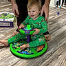 Zander joined the competition — help win amazing prizes! toddler, child, toy, green_pajamas, dinosaur, floor, adult_hands, baby_gate, smile, indoor, wood_flooring, playtime, barefoot, person, happy, furniture, box, colorful, support, home