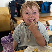 Zander is registered to the contest to win money with this photo: toddler, child, messy_face, food, bib, sippy_cup, plate, spoon, indoor, table, eating, happy, smiling, casual_clothing, hair, face, meal, mess, cute, person