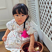 Lillian joined the competition — help win amazing prizes! toddler, child, girl, braid, pink_tassel, white_dress, floral_pattern, copper_bowl, flowers, wooden_floor, radiator_cover, indoor, curious, sitting, hand_in_bowl, natural_light, portrait, cute, decor, young_child