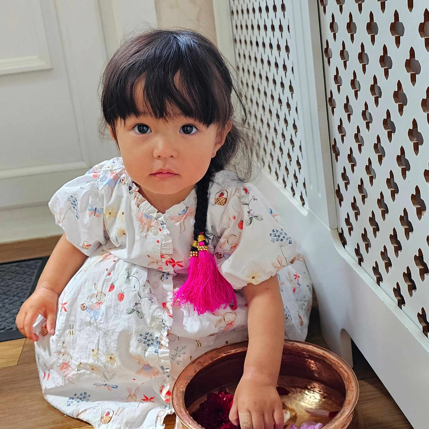 Lillian joined the competition — help win amazing prizes! braid, child, copper_bowl, curious, cute, decor, floral_pattern, flowers, girl, hand_in_bowl, indoor, natural_light, pink_tassel, portrait, radiator_cover, sitting, toddler, white_dress, wooden_floor, young_child