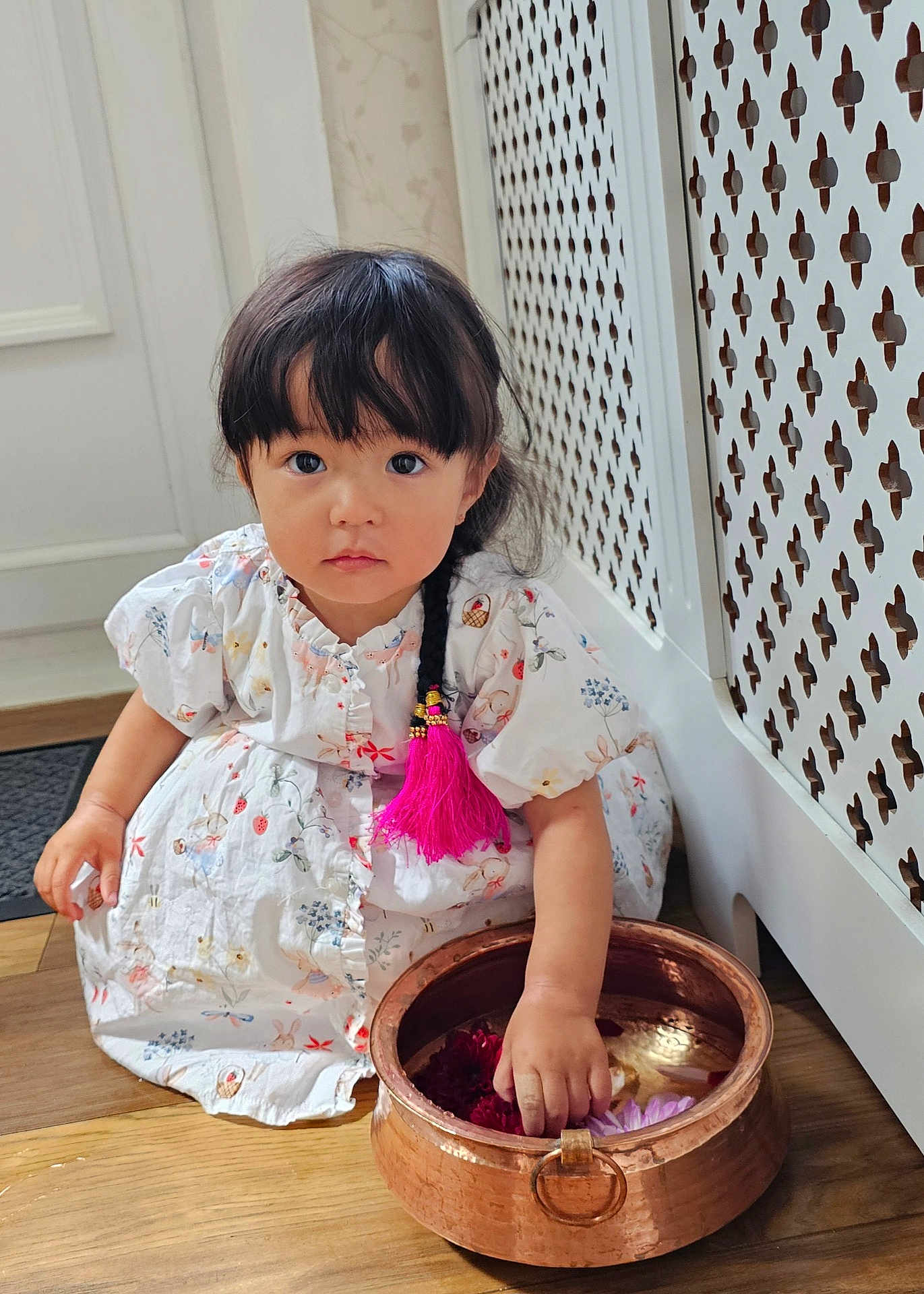 Lillian joined the competition — help win amazing prizes! toddler, child, girl, braid, pink_tassel, white_dress, floral_pattern, copper_bowl, flowers, wooden_floor, radiator_cover, indoor, curious, sitting, hand_in_bowl, natural_light, portrait, cute, decor, young_child