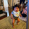 baby, child, infant, smiling, indoors, doorway_jumper, baby_bouncer, harness, dinosaur_bib, onesie, barefoot, family, adult, home, living_room, carpet, furniture, stroller, playtime, portrait