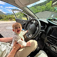 Albane a rejoint le concours — aidez-le/la à gagner de superbes lots ! baby, child, car_interior, steering_wheel, adult_hand, happy, smiling, road_trip, nature, trees, mountains, blue_sky, seat, clothing, person, sunlight, open_car_door, dashboard, comfort, playful