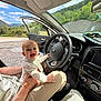 baby, child, car_interior, steering_wheel, adult_hand, happy, smiling, road_trip, nature, trees, mountains, blue_sky, seat, clothing, person, sunlight, open_car_door, dashboard, comfort, playful