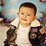 toddler, child, smile, jacket, aviator_jacket, patches, american_flag, indoor, portrait, face, hair, white_tshirt, cozy, background_blur, decorations, patriotic, cute, young_child, seated, casual_clothing