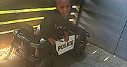 Major is registered to the contest to win money with this photo: black_shirt, boy, brick_wall, child, driveway, face, fence, outdoor, playtime, police_car, portrait, pretend_play, shadow, short_hair, sitting, smiling, sunlight, toddler, toy_vehicle, wheels