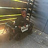 child, toddler, boy, police_car, toy_vehicle, outdoor, playtime, sitting, smiling, face, black_shirt, short_hair, driveway, fence, sunlight, shadow, brick_wall, wheels, pretend_play, portrait