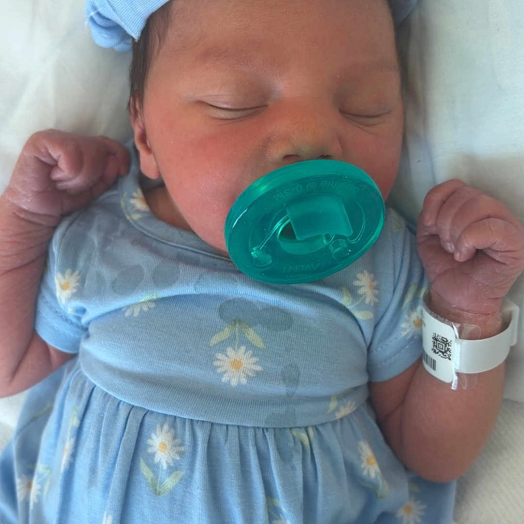 Tyanna is registered to the contest to win money with this photo: newborn, baby, sleeping, infant, pacifier, blue_dress, headband, bow, floral_pattern, hospital, wristband, portrait, close_up, lying_down, blanket, white_bedding, peaceful, hands, indoors, child