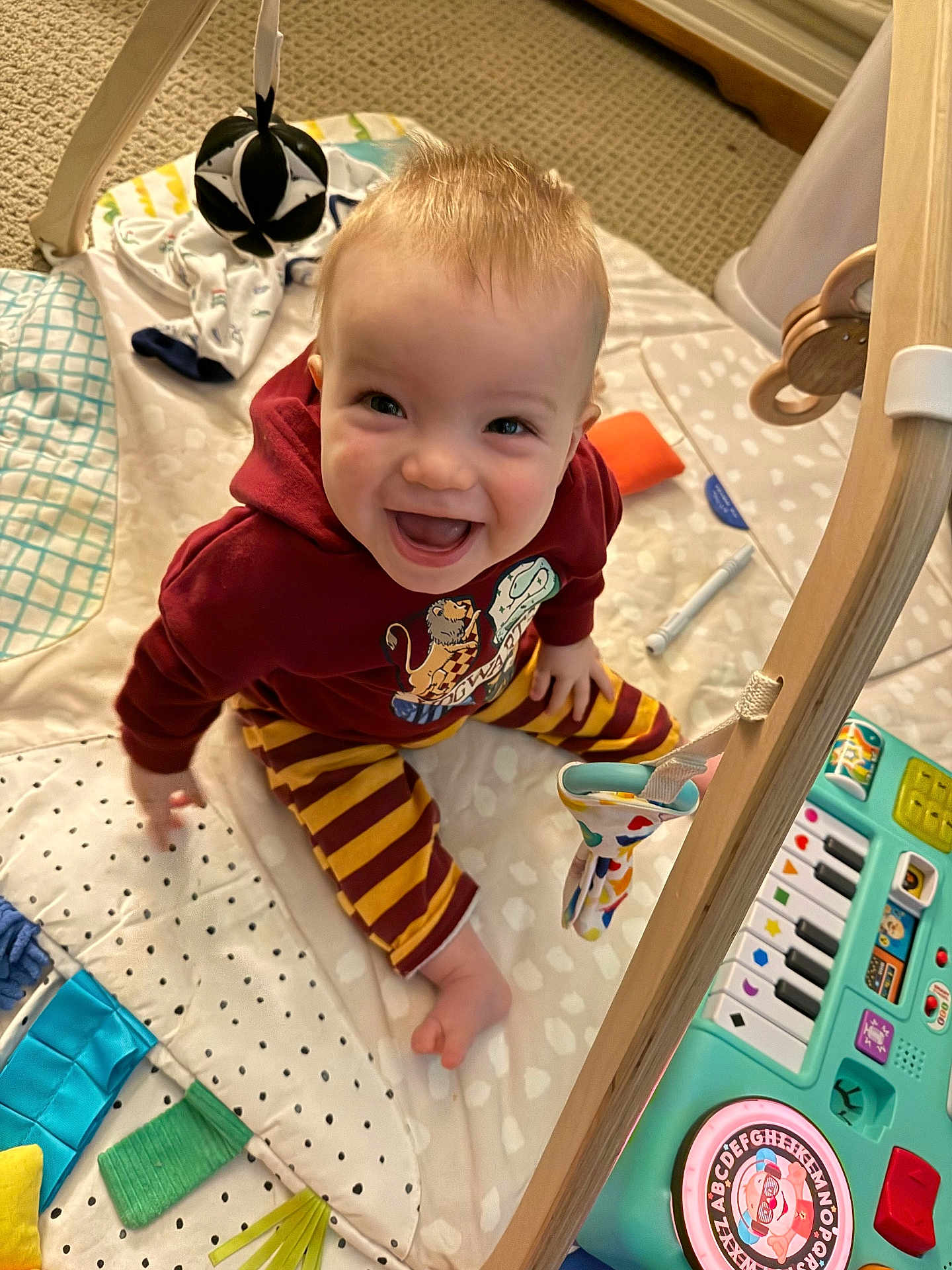 Archie is registered to the contest to win money with this photo: baby, child, smiling, sitting, play_mat, toys, hoodie, striped_pants, indoor, carpet, colorful, happy, cute, infant, playtime, person, face, young_child, floor, wooden_frame