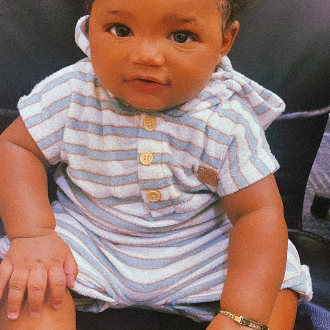 Journee Mcfadden is registered to the contest to win money with this photo: adorable, baby, bracelet, child, curious, cute, face, foot, hand, indoors, infant, leather_surface, person, pigtails, portrait, sitting, skin, smile, striped_clothing, young_child