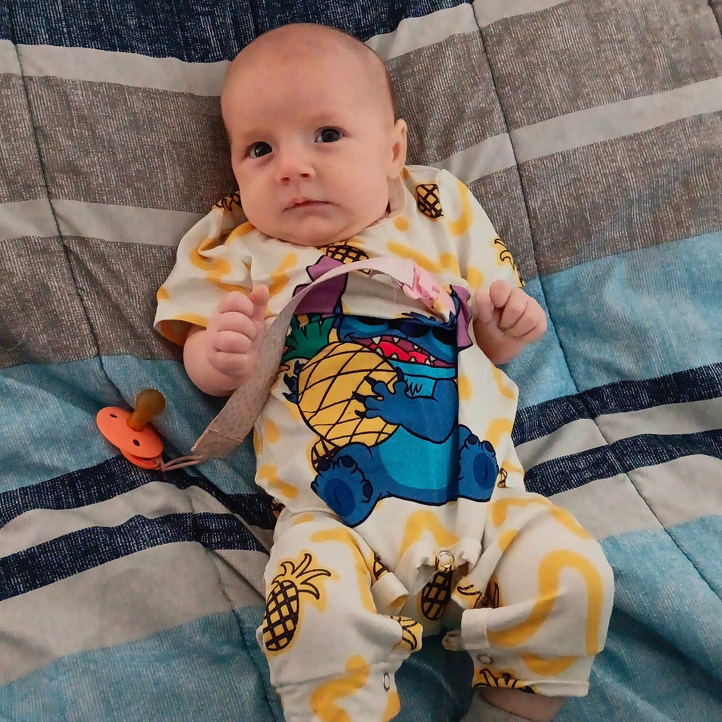 Fiona is registered to the contest to win money with this photo: baby, infant, onesie, pineapple, pacifier, blanket, striped, blue, gray, clothing, cute, child, person, sitting, indoor, soft, comfort, face, expression, sock