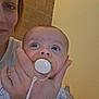 adult_hand, baby, brick_wall, closeup, clothing, comfort, expression, face, head, holding, human, indoor, infant, pacifier, parent, person, portrait, skin, yellow_wall, young_child