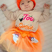 Nuyangma is registered to the contest to win money with this photo: baby, infant, tutu, orange_hat, pumpkin_hat, white_blanket, cute, lying_down, child, festive_clothing, shirt, text_on_shirt, soft_texture, indoors, sleepwear, costume, happy, smiling, portrait, toddler