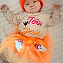 baby, infant, tutu, orange_hat, pumpkin_hat, white_blanket, cute, lying_down, child, festive_clothing, shirt, text_on_shirt, soft_texture, indoors, sleepwear, costume, happy, smiling, portrait, toddler