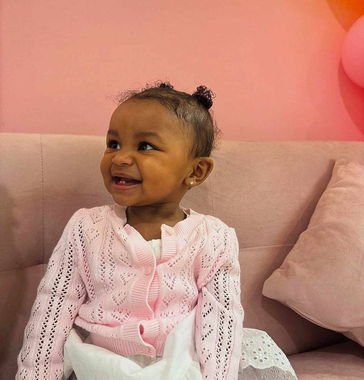 Ely joined the competition — help win amazing prizes! toddler, child, smile, pink_cardiagn, white_dress, curly_hair, earrings, couch, pillow, indoor, happy, cute, fashion, portrait, sitting, baby, cozy, soft_lighting, background, home