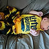 baby, bed, child, clothing, curious, cute, gray_bedspread, indoors, infant, lying_down, newborn, person, portrait, sibling, sleepwear, small_hands, soft_fabric, sunflower_pants, yellow_shirt, young_child
