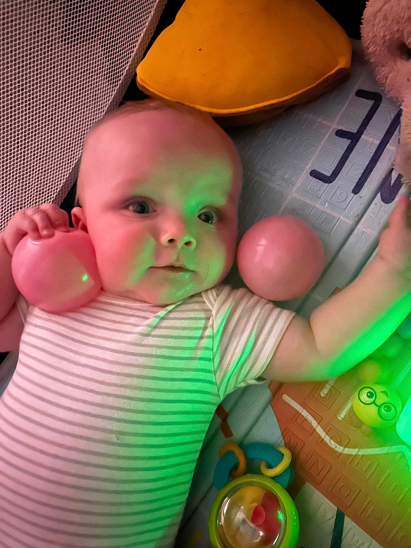 Oliver is registered to the contest to win money with this photo: baby, infant, play_mat, ball, pink_ball, onesie, striped_onesie, toys, rattle, pillow, plush_toy, mesh_side, green_light, close_up, face, eyes, hand, indoor, playtime, portrait