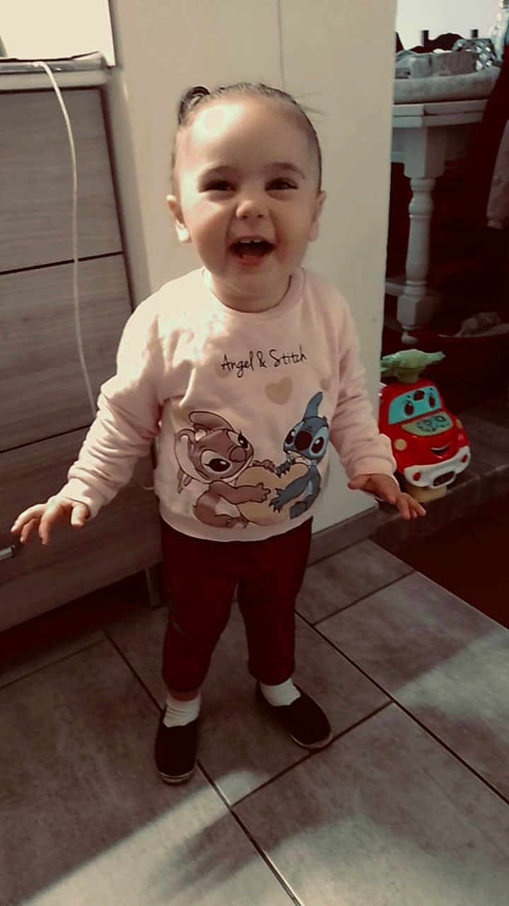 Lola participe au concours pour gagner de l'argent avec cette photo : toddler, child, laughing, standing, indoors, sweatshirt, angel_and_stitch, toy_car, floor, tile_floor, smile, person, clothing, happy, home, furniture, cabinet, socks, shoes, hair_tied