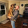 balloon, beard, bookshelf, brindle_dog, brown_dog, casual_clothing, curtains, dog, happy, holding, home_decor, indoor, living_room, man, plaid_shirt, plant, smiling, speaker, window, wooden_floor