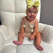 Brynli is registered to the contest to win money with this photo: baby, child, smiling, tongue_out, headband, bow, beige_outfit, flower, armchair, plush, textured, sitting, happy, indoor, cute, portrait, infant, person, skin, young_child