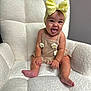 baby, child, smiling, tongue_out, headband, bow, beige_outfit, flower, armchair, plush, textured, sitting, happy, indoor, cute, portrait, infant, person, skin, young_child