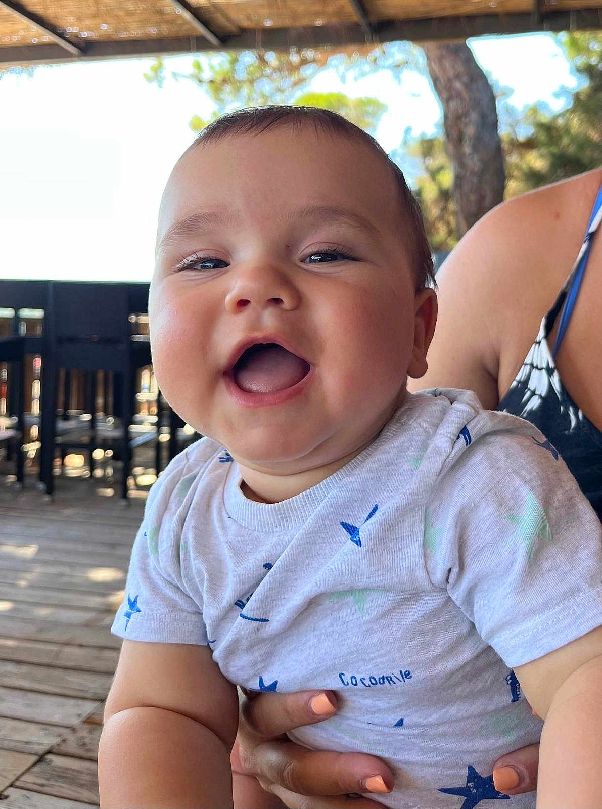 Pol a rejoint le concours — aidez-le/la à gagner de superbes lots ! baby, child, smiling, happy, person, face, shirt, pattern, outdoor, wooden_floor, hand, holding, seat, table, tree, nature, sunlight, shade, casual, portrait