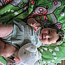 baby, infant, child, smiling, portrait, lying_down, blanket, green_blanket, teddy_bear, stuffed_animal, plush_toy, headband, bow, knitted_clothing, dress, bare_legs, hand, fist, indoor, cozy